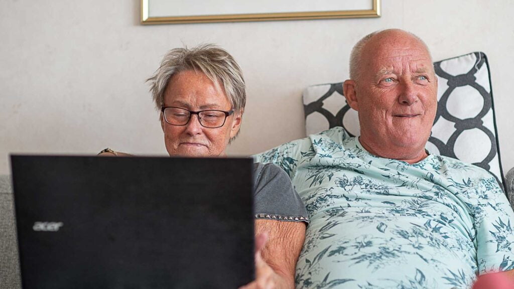 Två seniorer sitter i soffa med en bärbar dator framför sig.