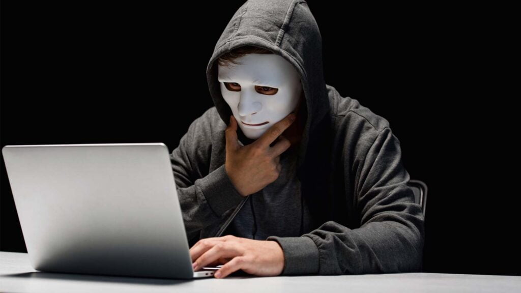Person som döljer sitt ansikte bakom en mask sitter framför sin dator.