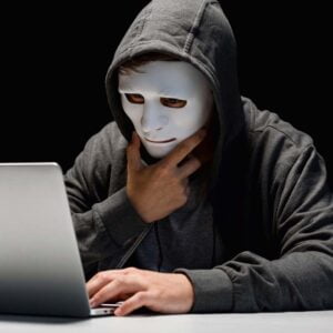 Person som döljer sitt ansikte bakom en mask sitter framför sin dator.