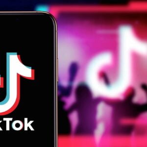 En mobil visar Tiktoks logotyp. I bakgrunden syns personer dansa i en färgsprakande lokal med Tiktoks logotyp på väggen.