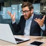En upprörd man sitter vid sin laptop i en kontorsmiljö.