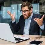 En upprörd man sitter vid sin laptop i en kontorsmiljö.